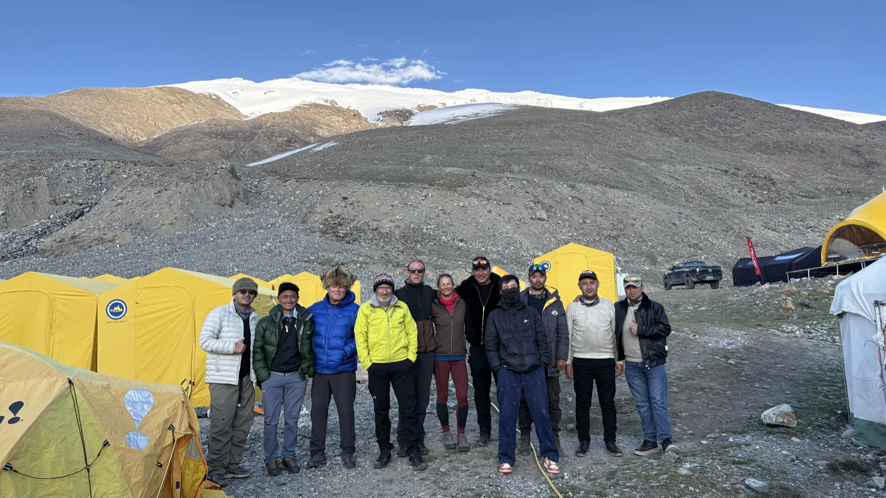 Climbers and camp staff standing together among yellow expedition tents beneath Muztagh Ata at Base Camp.