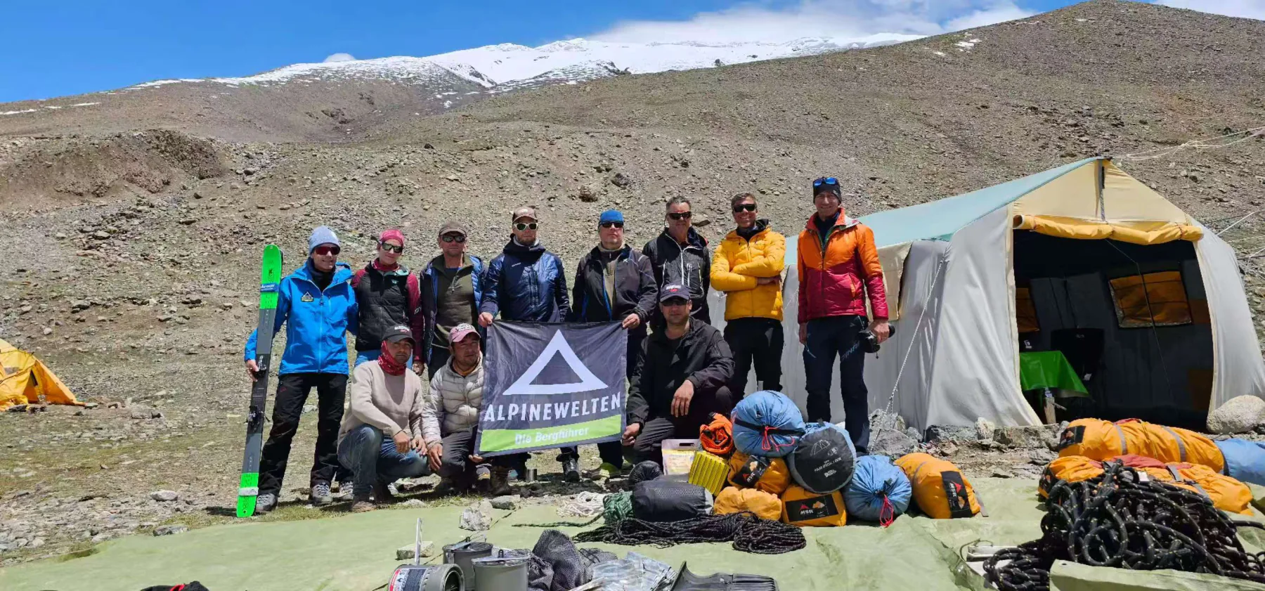 An expedition team posing with skis, packed duffels, ropes, and a team flag beside the Base Camp dining tent.