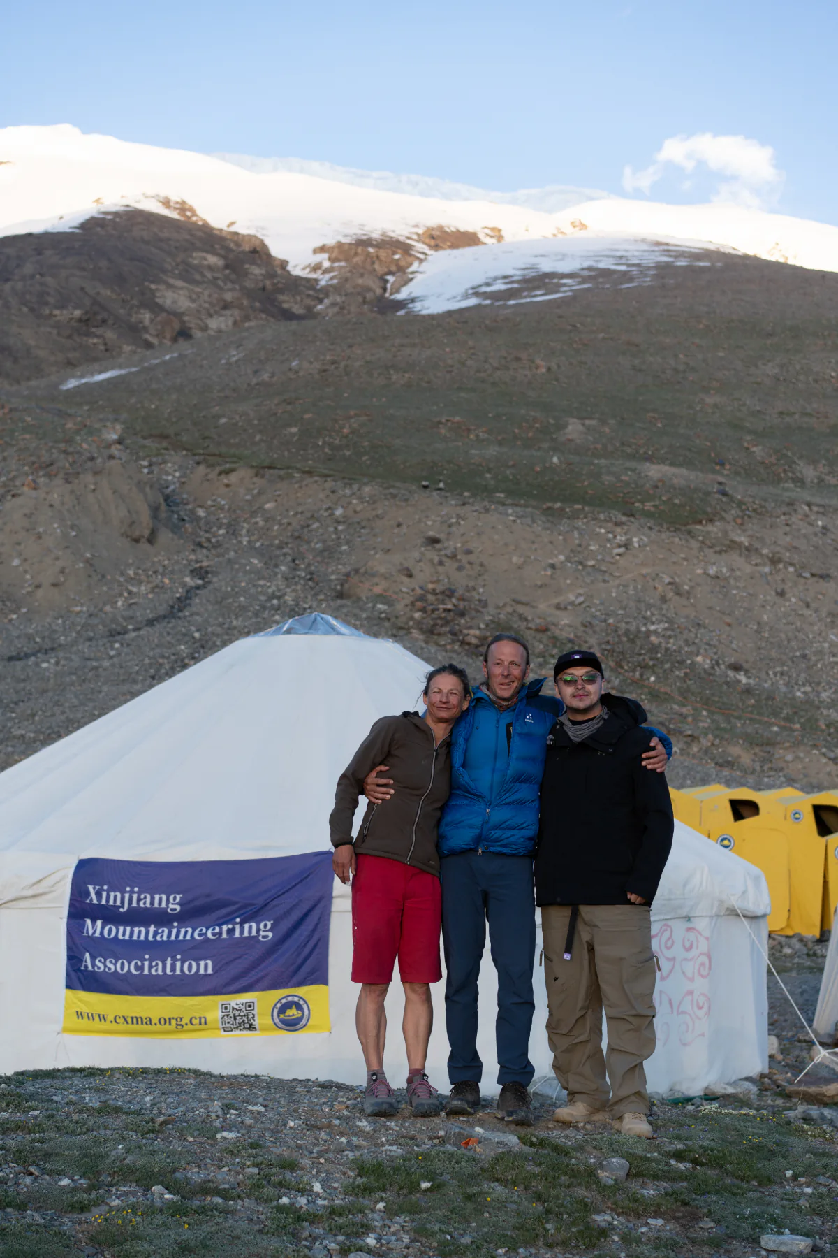 Three expedition members standing in front of a CXMA communal tent with the snow-covered mountain above.