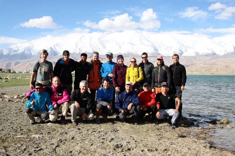 An expedition team standing beside Karakul Lake with the snowy Muztagh Ata massif behind.