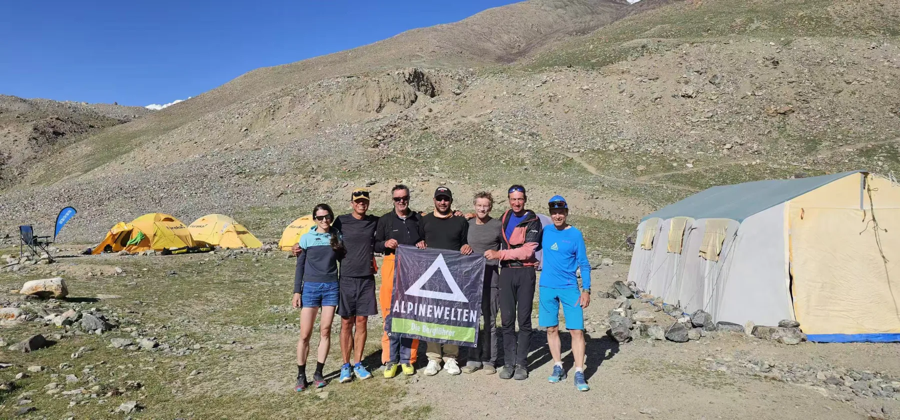 Seven expedition members holding a team flag between sleeping tents and the Base Camp dining tent.