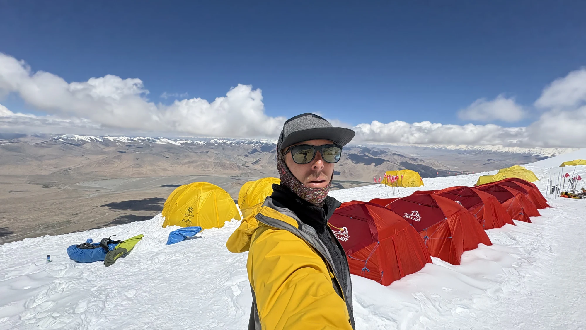 A climber standing among red and yellow expedition tents on the snow at Camp 1, with the Pamir landscape below.