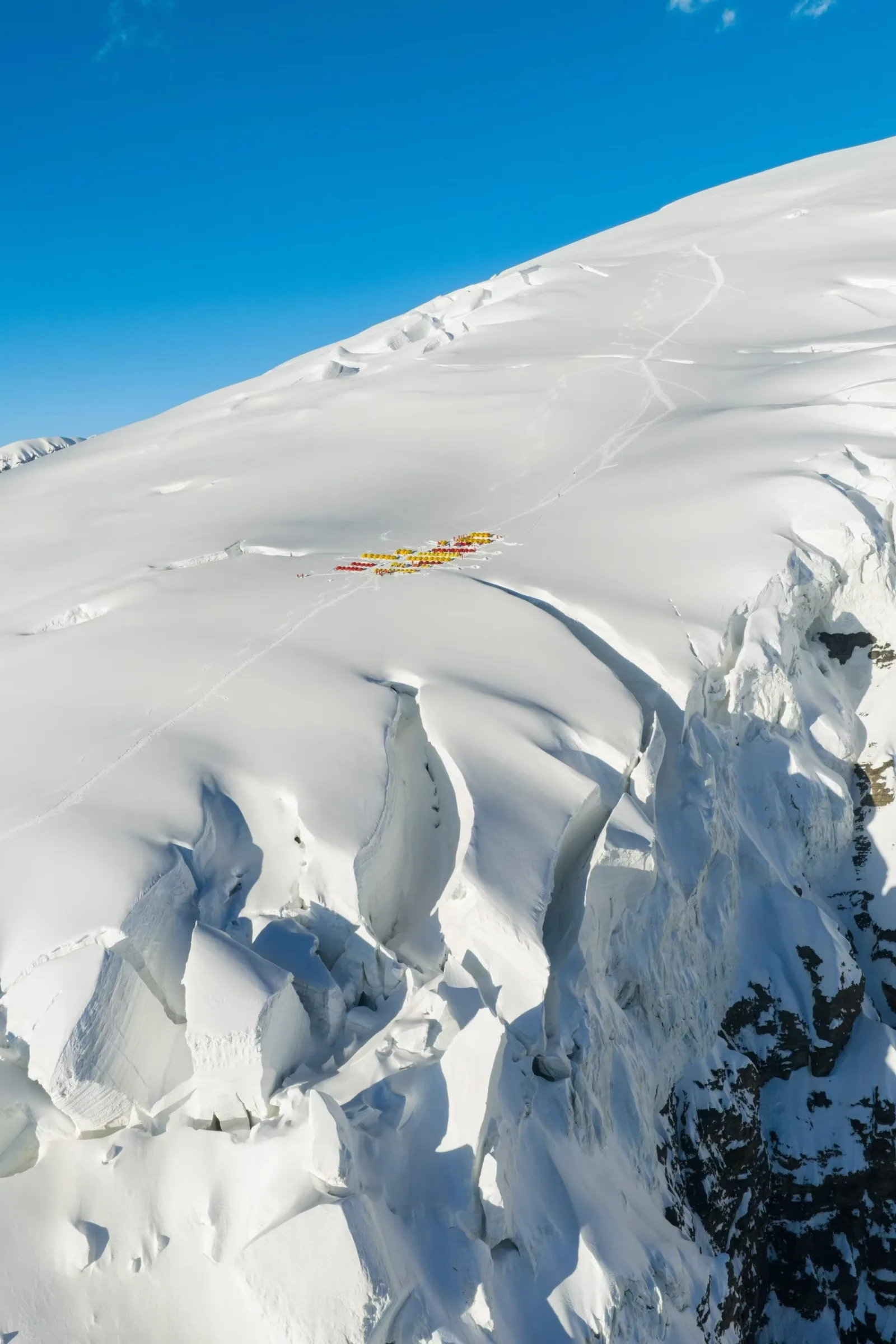 Red and yellow expedition tents at Camp 2 on a small glacier shelf surrounded by crevasses and steep snow terrain.