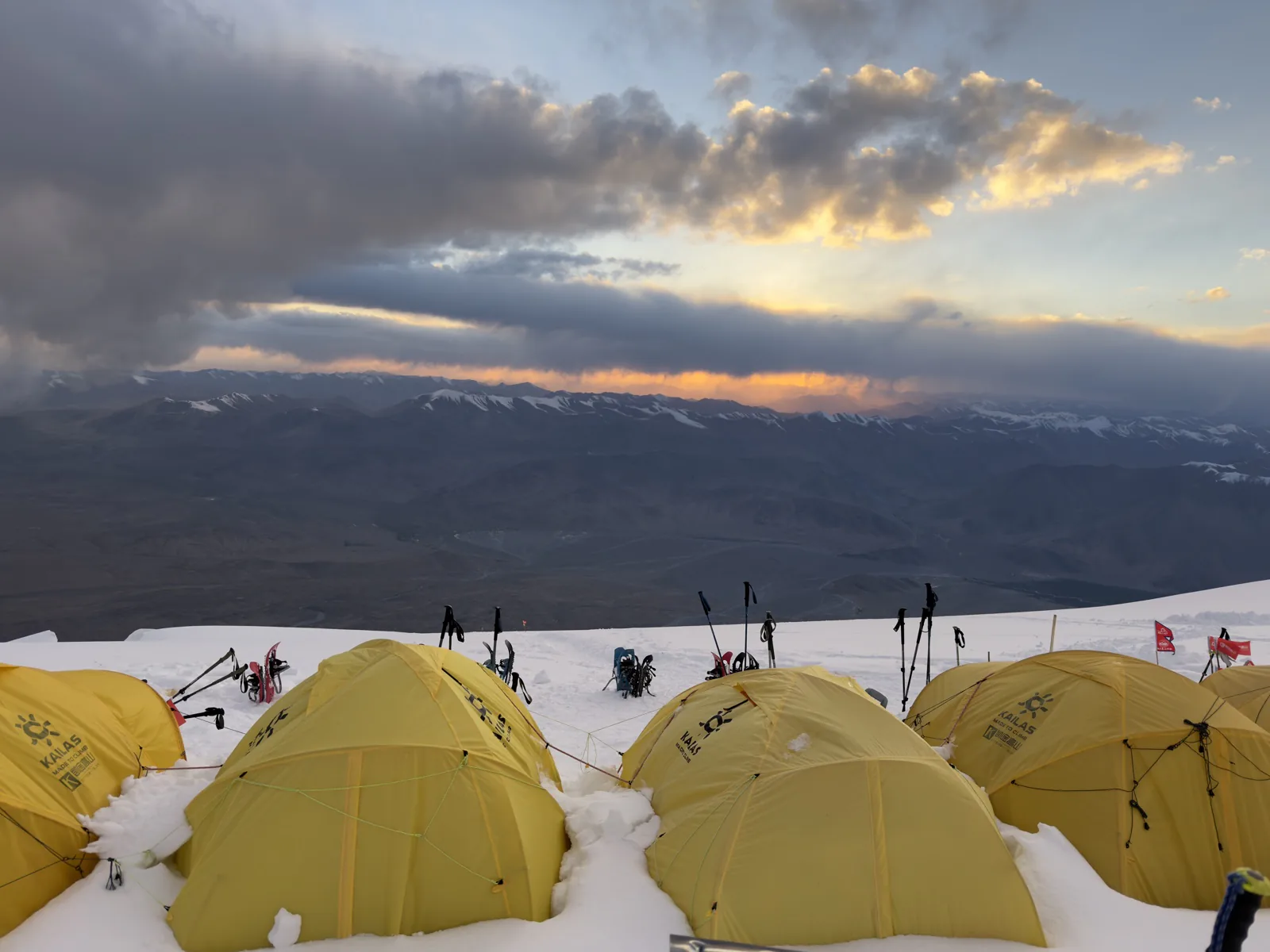 Yellow expedition tents on snow above the Pamir range beneath dramatic evening clouds.