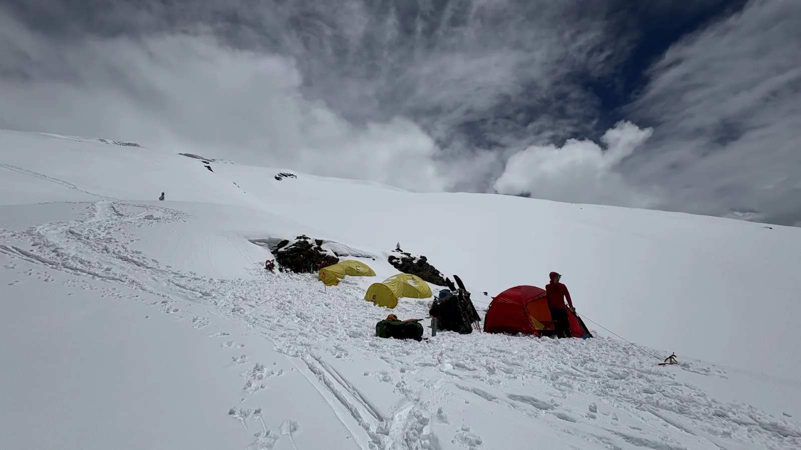A small group of yellow and red expedition tents cut into a steep snow slope.