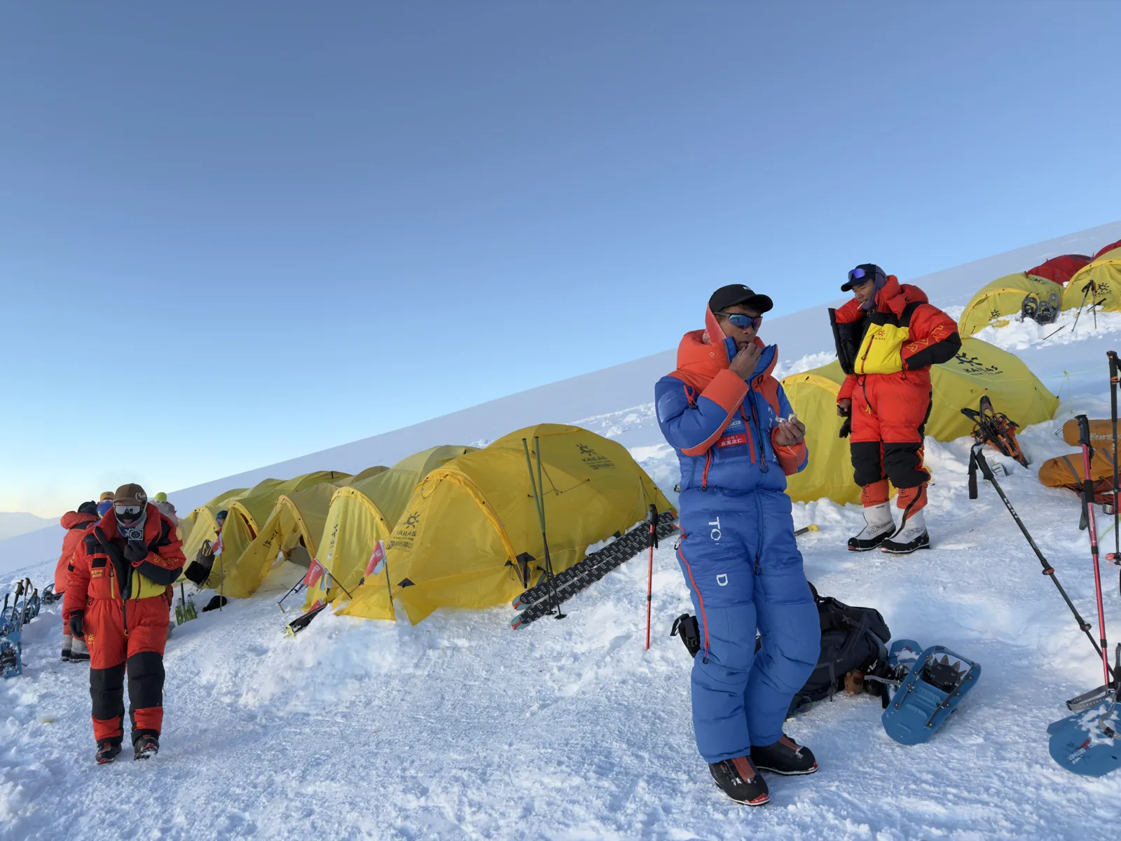 Climbers in high-altitude suits preparing equipment among yellow tents on the snow.