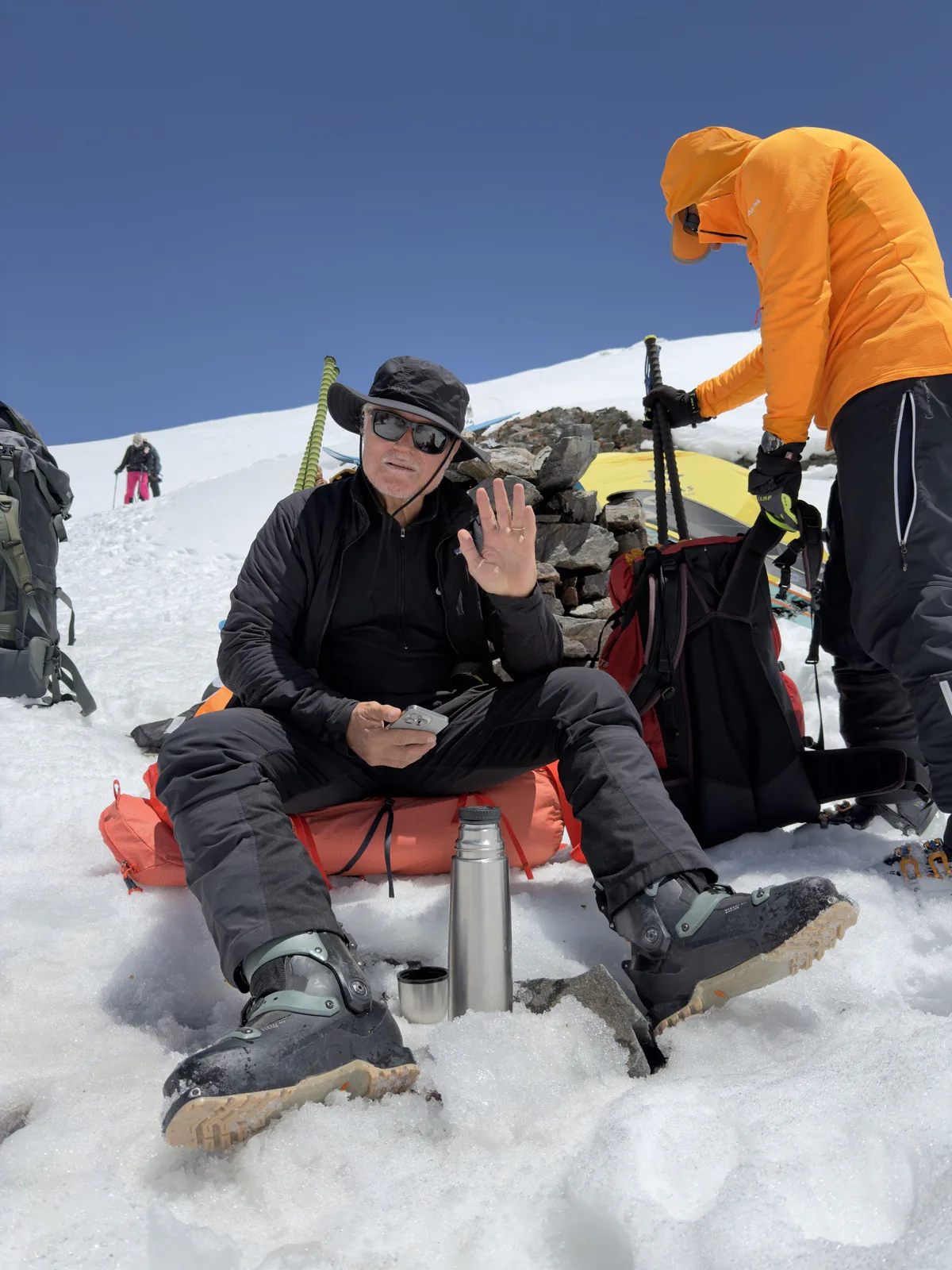 A climber resting on the snow with a flask and expedition equipment beside camp.