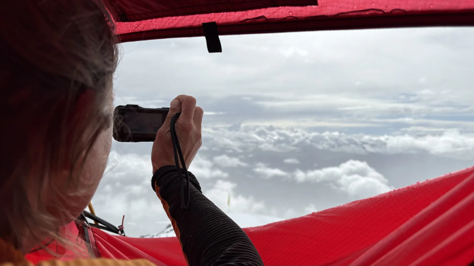 A climber photographing a sea of clouds from inside a red expedition tent.