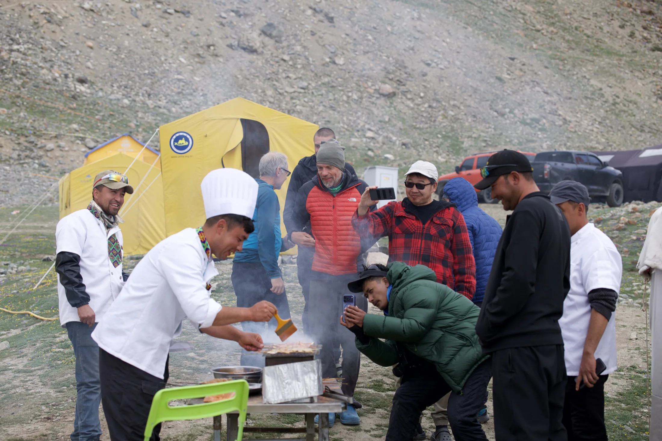 Camp cooks grilling food while climbers and staff gather beside yellow expedition tents at Base Camp.