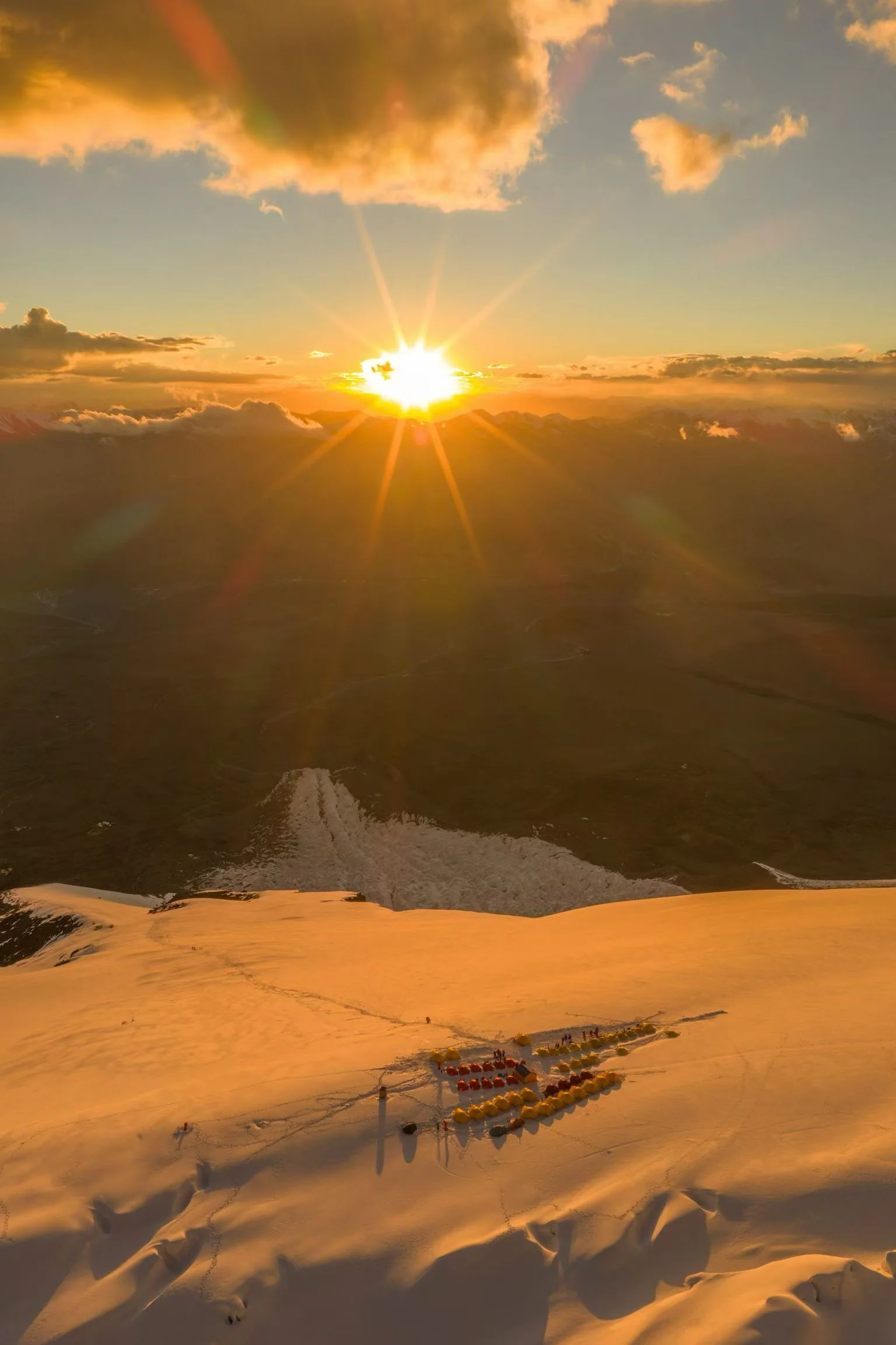 A high-altitude tent camp glowing in golden sunset light beneath the mountain horizon.