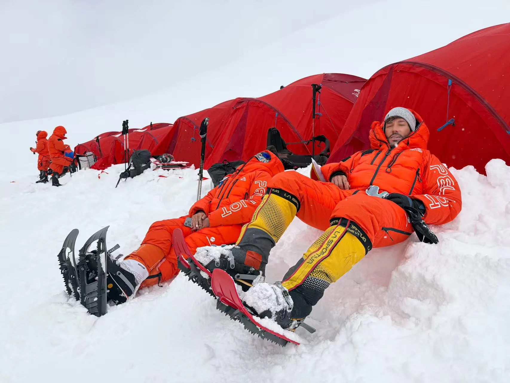 Two climbers in orange high-altitude suits resting on the snow beside red expedition tents.
