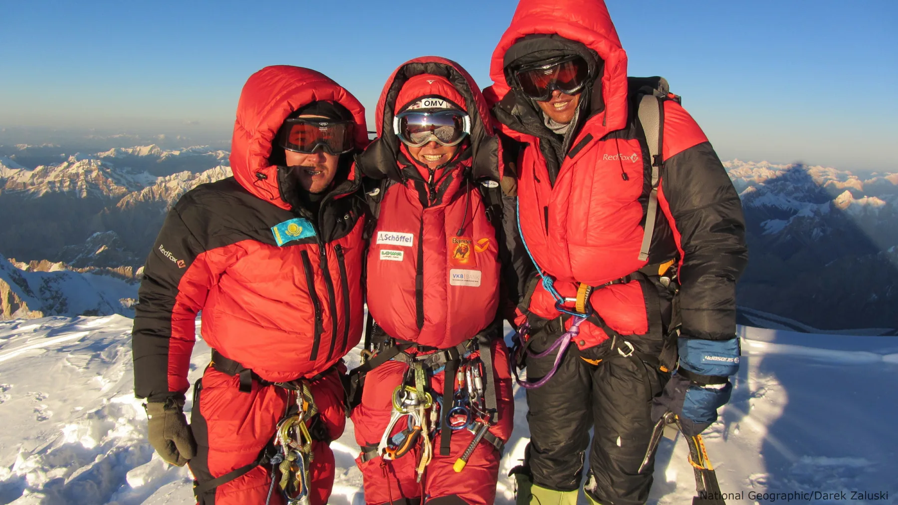 Three climbers in red high-altitude suits standing shoulder to shoulder on a snowy summit above a mountain range.