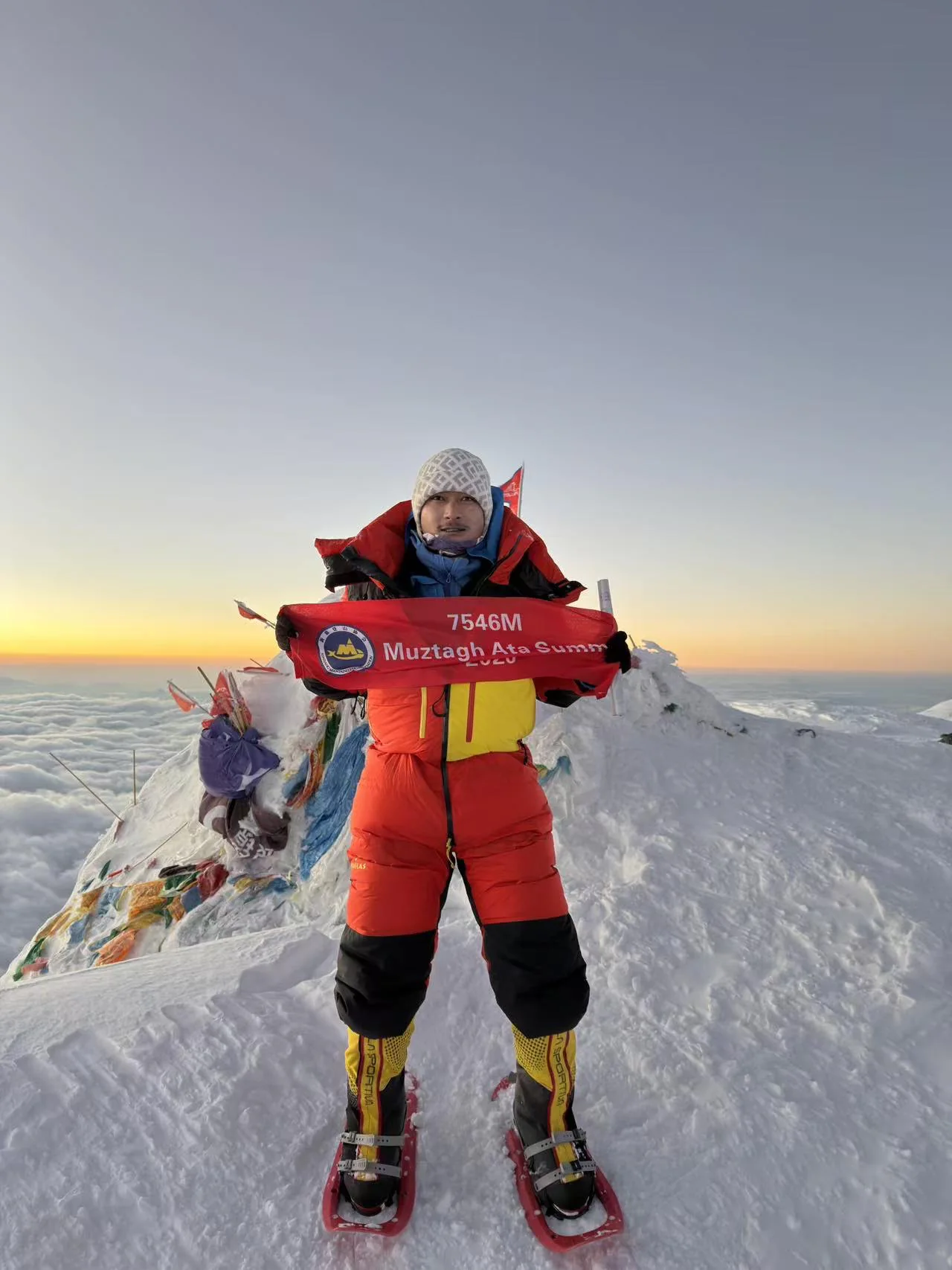 A climber in a red high-altitude suit holding a 7,546-metre Muztagh Ata summit banner at sunrise above the clouds.