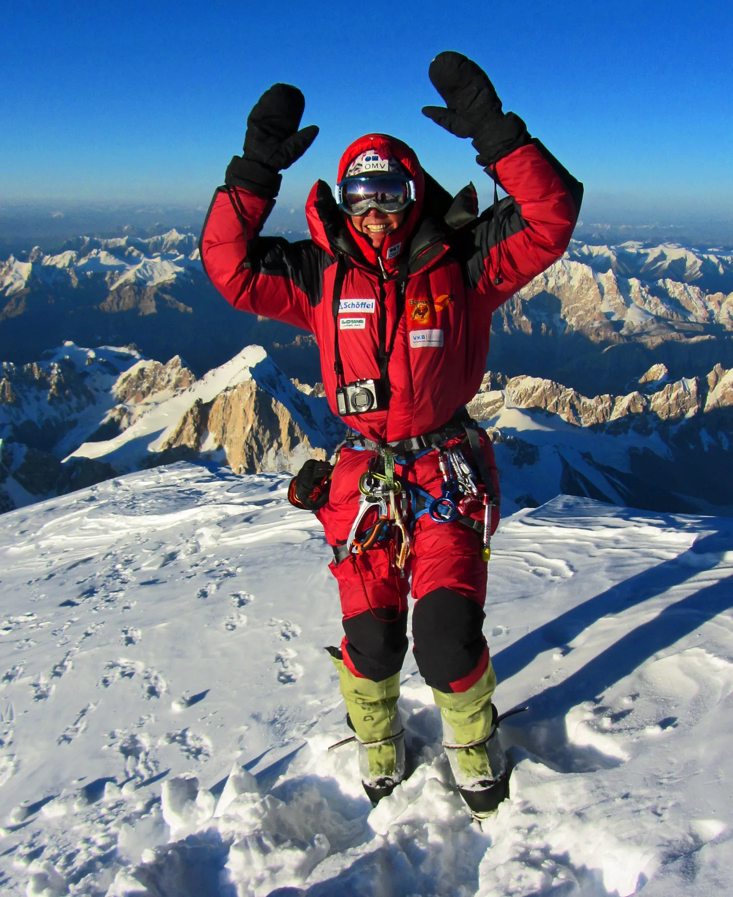 A climber in a red high-altitude suit raising both arms on a snowy summit with mountain ranges behind.