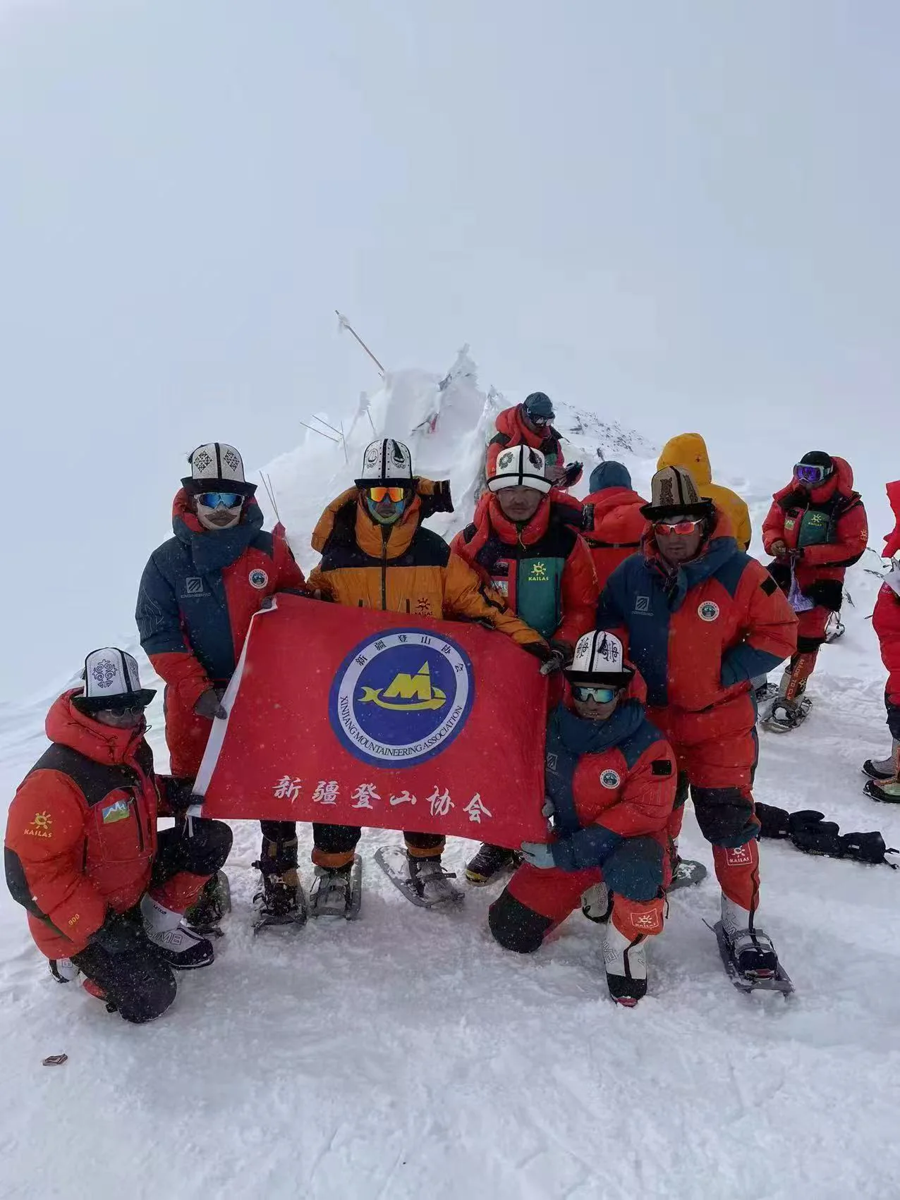 A group of climbers in winter gear holding the Xinjiang Mountaineering Association flag in snowy summit conditions.