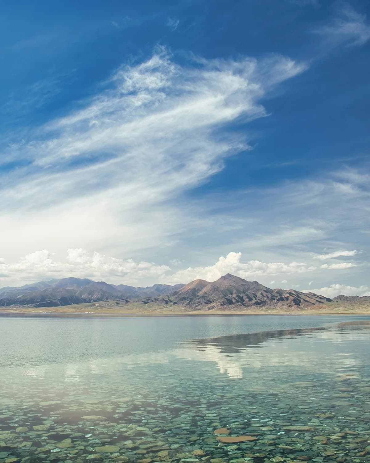 Stones visible through clear lake water below a distant mountain ridge and blue sky.