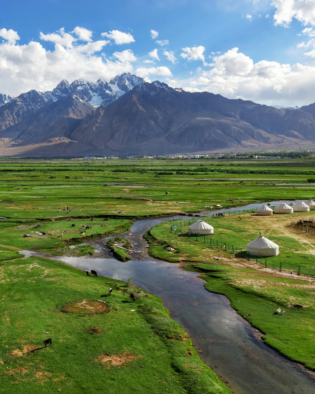 A stream winding through green pasture towards yurts and distant mountain peaks.