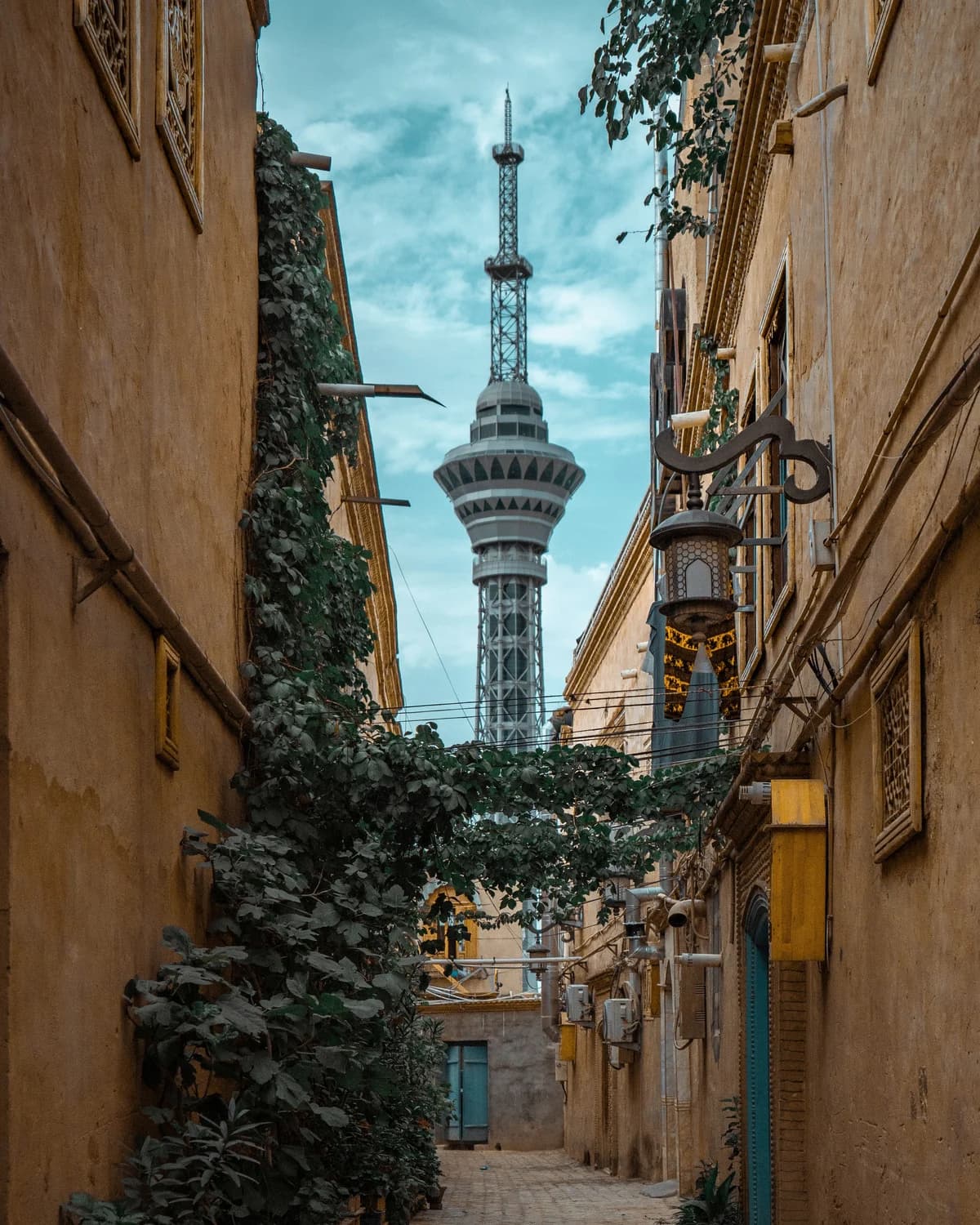 The Kashgar TV tower rising between ochre walls above a narrow alley with climbing plants.