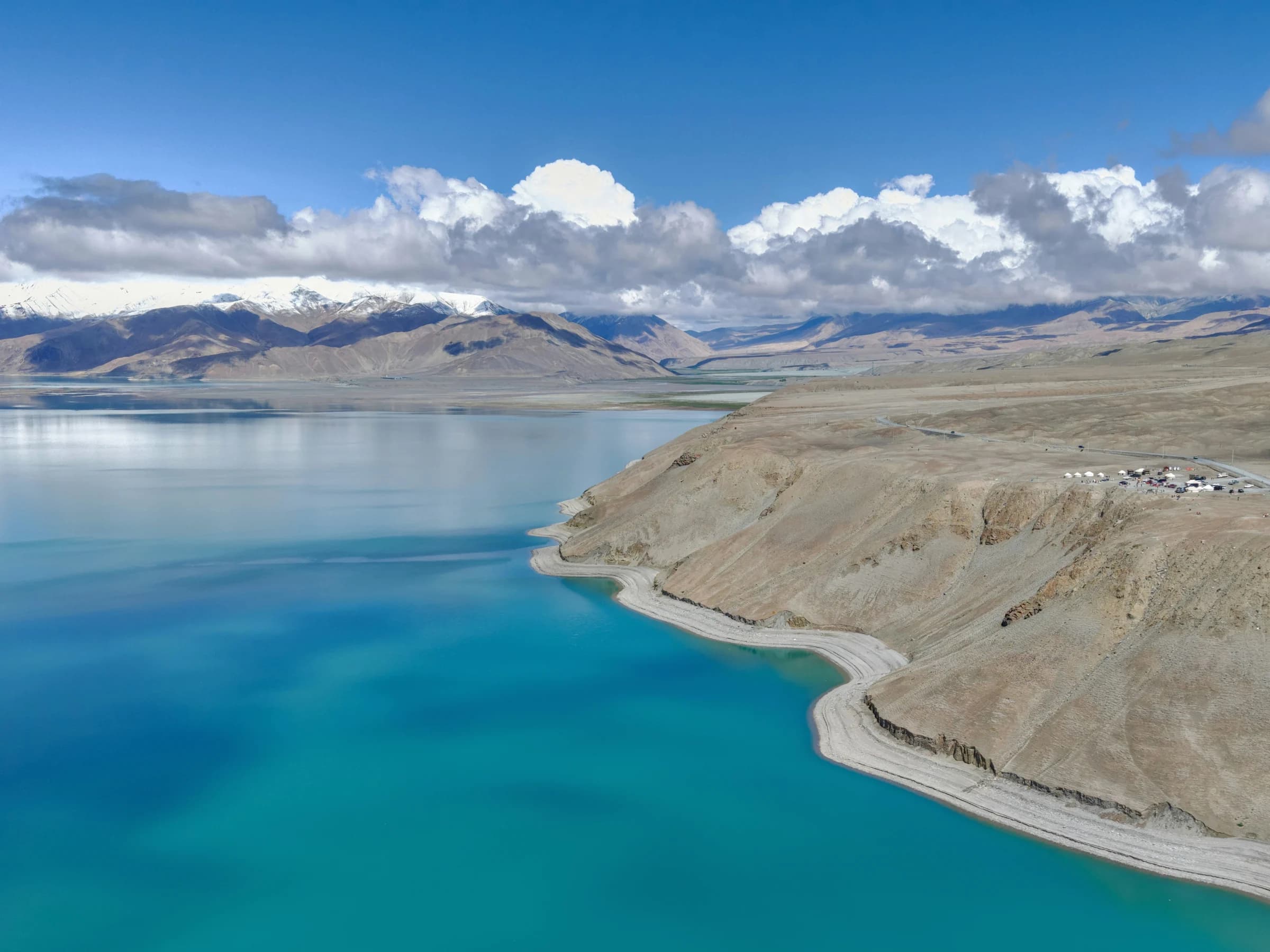 A pale shore beside turquoise lake water with arid slopes and snow-covered mountains beyond.