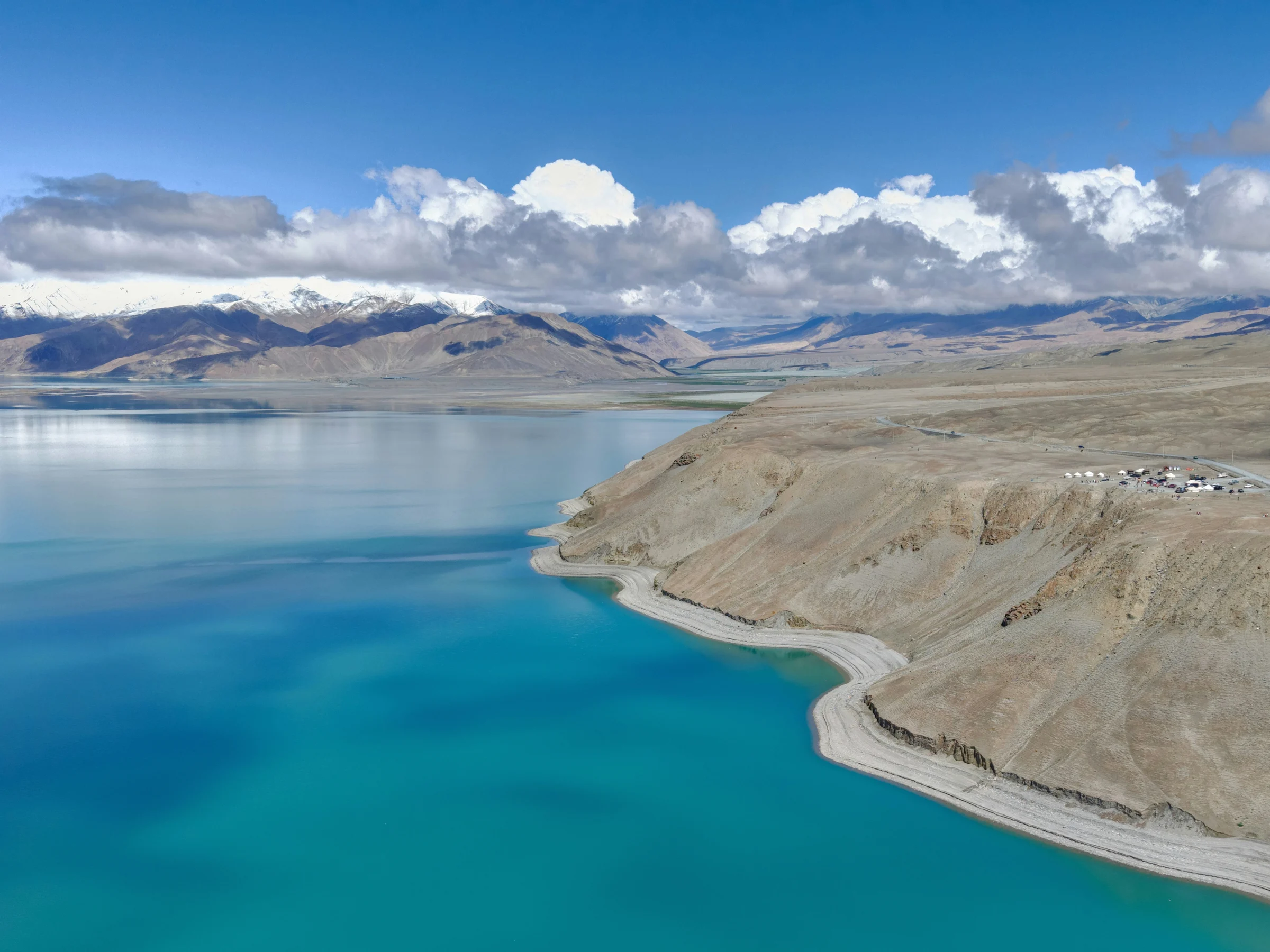 A pale shore beside turquoise lake water with arid slopes and snow-covered mountains beyond.